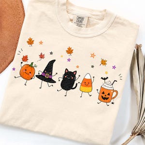 Peut inclure: Un t-shirt couleur crème avec une étiquette Comfort Colors présentant une impression graphique d'une citrouille souriante, un chapeau de sorcière, un chat noir, un bonbon maïs et une tasse jack-o'-lantern avec de la crème fouettée.