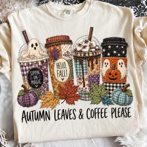 Puede incluir: Una camiseta color crema con un gráfico de cuatro tazas de café decoradas con motivos de otoño y Halloween, incluyendo fantasmas, calabazas y hojas. El texto dice "Sippin' on Magic," "Hello, Fall!," y "Autumn Leaves & Coffee Please."