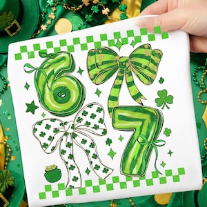 Puede incluir: Camiseta blanca con los números "67" en verde, adornada con lazos. El "6" tiene un diseño en espiral, mientras que el "7" es rayado. Los lazos tienen detalles dorados y patrones de tréboles. También hay un pote de oro y tréboles.