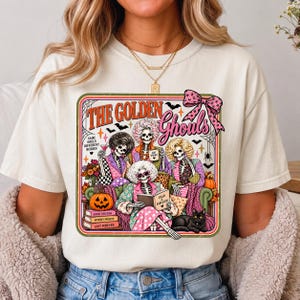 Puede incluir: Una camiseta de color crema con un estampado gráfico de cuatro esqueletos con peinados elaborados sentados en un sofá. El texto dice THE GOLDEN GHOULS, SAME SOULS DIFFERENT BODIES, GOOD FRIENDS, SPOOKY NIGHTS, LAST FOREVER y DEAD AND FABULOUS.