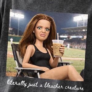 Puede incluir: Camiseta gris oscuro con un gráfico de una muñeca con cabello castaño rojizo, que lleva una camiseta sin mangas negra y sostiene un café helado. La muñeca está sentada en una silla plegable con un estadio de béisbol al fondo. El texto "literally just a bleacher creature" está en la parte inferior.