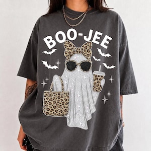 Puede incluir: Una camiseta gris carbón de manga corta con el gráfico de un fantasma con gafas de sol y un lazo de estampado de leopardo. El fantasma sostiene un bolso y un vaso con estampado de leopardo. El texto BOO-JEE aparece arriba con murciélagos blancos.