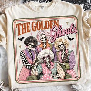 Puede incluir: Una camiseta color crema con un gráfico de cuatro esqueletos con peinados elaborados y blazers estampados de colores. El texto dice THE GOLDEN Ghouls, acompañado de pequeñas ilustraciones de estrellas y murciélagos en un borde rectangular.