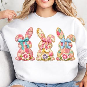 Puede incluir: Sudadera blanca con tres diseños de conejitos de Pascua aplicados. Cada conejito está hecho de tela de patchwork en rosas, amarillos y azules, con lazos y adornos florales. Un diseño festivo y colorido para la temporada de primavera.