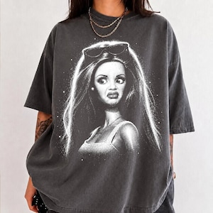 Puede incluir: Una camiseta gris carbón de corte oversize con un estampado gráfico blanco de un personaje estilizado de pelo largo, con gafas de sol sobre la cabeza y una camiseta de tirantes. El diseño incluye sutiles motas blancas alrededor.