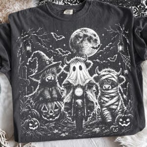 Puede incluir: Una camiseta gris carbón con un estampado gráfico blanco de tres vacas Highland con disfraces de Halloween. Una vaca lleva un sombrero de bruja sobre una calabaza, otra es un fantasma en una motocicleta y la tercera es una momia.