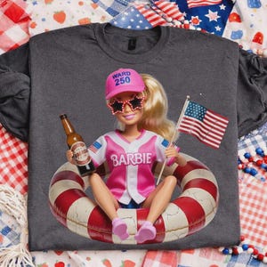 May include: Dark gray t-shirt featuring a Barbie doll in a pink baseball jersey, star-shaped sunglasses, and a pink cap that says "WARD 250." The doll is holding a beer bottle and an American flag while sitting in a red and white striped inner tube.