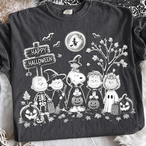 May include: A charcoal gray t-shirt featuring a white graphic of Peanuts characters in Halloween costumes. The design includes a wooden sign reading Happy Halloween, a full moon with a witch silhouette, bats, pumpkins, and falling leaves.