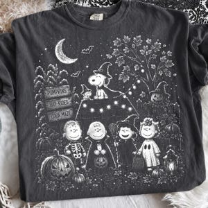 May include: A charcoal gray t-shirt featuring a white graphic print of Peanuts characters in Halloween costumes. The design includes Snoopy on a doghouse, a sign reading PUMPKINS, HAY-RIDES, CORN MAZE, a crescent moon, bats, and pumpkins.
