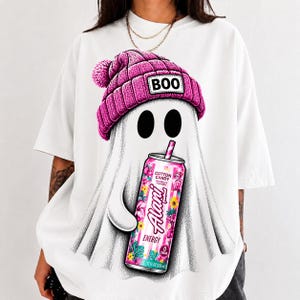 Op de afbeelding: Een wit t-shirt met een afbeelding van een geest die een roze gebreide muts met pompon en de tekst BOO draagt. De geest houdt een roze Alani energiedrankblikje vast met bloemmotieven en de tekst Cotton Candy, 12 FL OZ 355 ml.