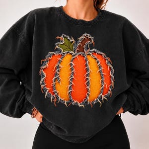 Puede incluir: Una sudadera de cuello redondo en color negro carbón con un gran gráfico central de calabaza. La calabaza está construida con segmentos de tela superpuestos de bordes deshilachados en tonos naranja y amarillo, con una hoja verde y un tallo marrón.