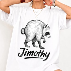 May include: A white short-sleeved t-shirt featuring a black and white line art illustration of a raccoon walking. Below the raccoon, the name Jimothy is printed in a bold, black, handwritten-style script font across the center of the chest.
