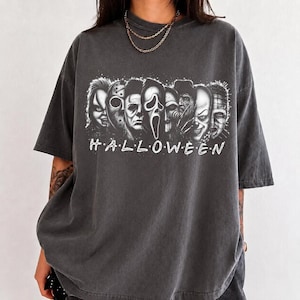 May include: A charcoal grey short-sleeved t-shirt featuring a black and white graphic print of various horror movie characters arranged in a row. Below the characters, the word HALLOWEEN is printed in a white, stylised, sans-serif font.
