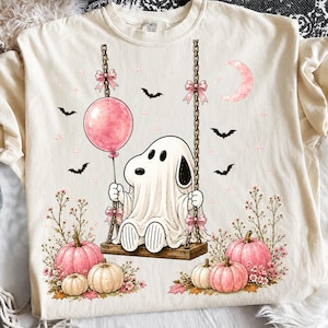 Puede incluir: Una camiseta de color crema con un gráfico de un personaje fantasma blanco sentado en un columpio de madera. El fantasma sostiene un globo rosa y está rodeado de calabazas rosas, flores pequeñas, murciélagos y una luna creciente rosa. El columpio cuelga de cadenas decoradas con lazos rosas.