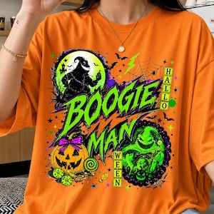 Puede incluir: Una camiseta naranja con un estampado gráfico que incluye el texto "BOOGIE MAN" y "HALLOWEEN". El diseño muestra la silueta de un personaje frente a una luna amarilla, una calabaza con un lazo morado, dados verdes y un personaje verde.