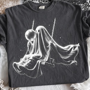 Puede incluir: Una camiseta gris carbón con una ilustración gráfica blanca de un fantasma sentado en un columpio, sosteniendo un orbe brillante. El fantasma tiene apariencia de sábana, rodeado de pequeñas estrellas, con la etiqueta Comfort Colors visible.