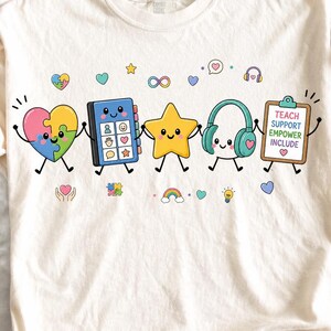 Puede incluir: Una camiseta de color crema con un gráfico de cinco personajes de dibujos animados tomados de la mano: un corazón de piezas de rompecabezas, un tablero de comunicación azul, una estrella amarilla, auriculares y un portapapeles con el texto TEACH SUPPORT EMPOWER INCLUDE.