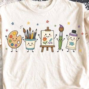 Puede incluir: Una camiseta de color crema con un estampado gráfico de cinco artículos de arte antropomórficos, incluyendo una paleta de pintura, un vaso con pinceles y lápices, un caballete con un lienzo, un pincel y un tubo de pintura, rodeados de estrellas.