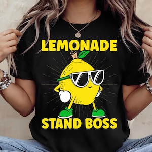 May include: Black t-shirt featuring a cartoon lemon wearing sunglasses and green shoes. The text "LEMONADE STAND BOSS" is printed in yellow. The lemon has a green headband and a green leaf on top.