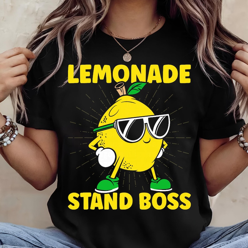 May include: Black t-shirt featuring a cartoon lemon wearing sunglasses and green shoes. The text "LEMONADE STAND BOSS" is printed in yellow. The lemon has a green headband and a green leaf on top.