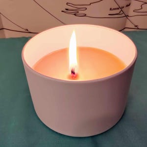 Könnte beinhalten: Eine brennende Kerze in einem weißen, zylindrischen Behälter. Die Kerze hat ein warmes, orange-gelbes Wachs und eine helle, flackernde Flamme. Der Behälter steht auf einer türkisfarbenen Oberfläche, mit einer Karte im Hintergrund.