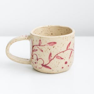 May include: A beige ceramic mug with a rounded handle and a floral design. The mug features a speckled texture and is adorned with a red floral pattern. The design includes flowers and leaves.