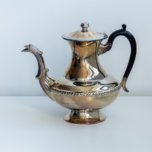 May include: A silver-toned teapot with a black handle and a decorative spout. The teapot has a bulbous body and a lid with a small knob. The design is ornate, with detailed patterns around the base and lid. The teapot is likely made of metal.