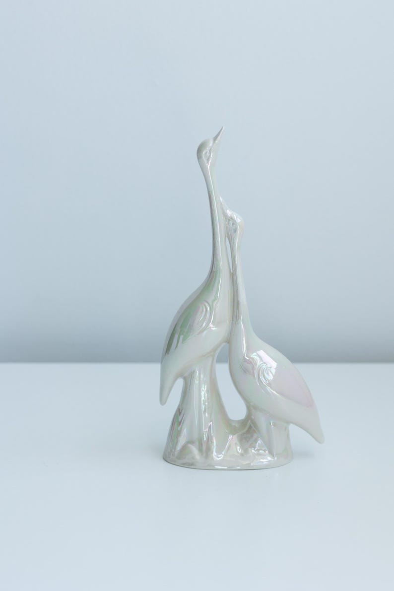 Może przedstawiać: Perłowa ceramiczna figurka przedstawiająca dwa stylizowane żurawie. Ptaki są splecione, z długimi szyjami i delikatnymi skrzydłami. Rzeźba ma błyszczące wykończenie i stoi na białej powierzchni.
