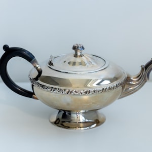 May include: A silver teapot with a black handle and spout. The teapot features a decorative floral pattern around the base and lid. The handle is curved and the spout is shaped like a dragon's head. The teapot is on a white surface.