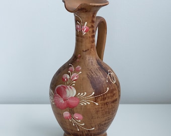 Op de afbeelding: Een bruine keramische kan met een handvat en een brede tuit. De kan is versierd met roze en witte bloemmotieven. De basis heeft een wit zigzagpatroon. De kan is ongeveer 25 cm hoog.