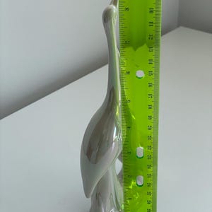 Może przedstawiać: Perłowa ceramiczna figurka żurawia, o wysokości około 28 cm, stoi wdzięcznie. Rzeźba ma długą szyję i zakrzywione ciało, z gładkim, błyszczącym wykończeniem. Figurka jest przedmiotem dekoracyjnym.