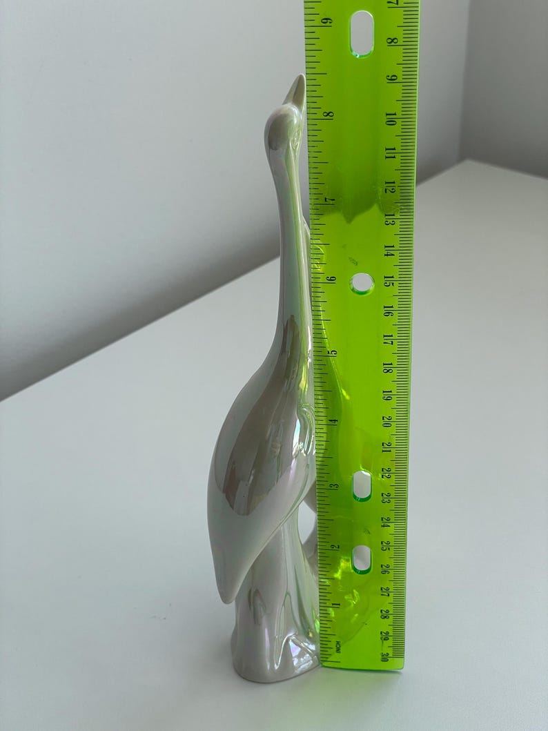 Może przedstawiać: Perłowa ceramiczna figurka żurawia, o wysokości około 28 cm, stoi wdzięcznie. Rzeźba ma długą szyję i zakrzywione ciało, z gładkim, błyszczącym wykończeniem. Figurka jest przedmiotem dekoracyjnym.