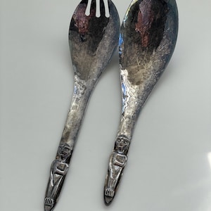 May include: A pair of vintage silver salad servers. One server is a fork, the other a spoon. Each handle features a detailed figure. The servers have a tarnished finish, giving them a classic, antique look. The set is designed for serving salads.