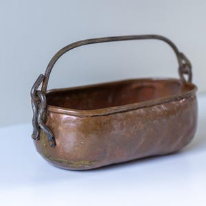 May include: An oval-shaped copper vessel with a hammered texture and a dark, weathered patina. The container features a sturdy, arched iron handle attached to the sides with metal loops, showcasing a rustic appearance with visible signs of age.