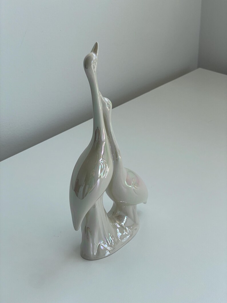 Może przedstawiać: Perłowa, biała ceramiczna figurka dw&oacute;ch stylizowanych ptak&oacute;w, być może żurawi lub czapli, stojących blisko siebie. Ptaki mają długie szyje i siedzą na ozdobnej podstawie. Figurka ma błyszczące wykończenie.
