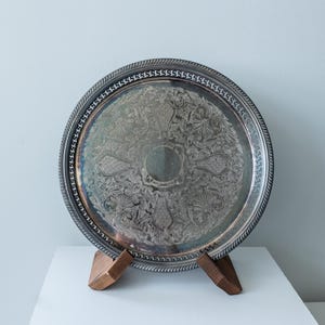 Puede incluir: Bandeja plateada con un diseño ornamental de flores y volutas. La bandeja tiene un borde detallado en forma de cuerda y se exhibe sobre un soporte de madera. Una pieza decorativa para servir o exhibir.