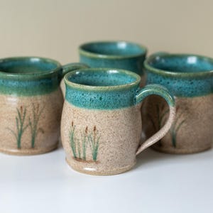 May include: A set of four ceramic mugs with a speckled tan base and a teal glazed rim. Each mug features a hand-painted design of green cattail plants on the lower body. The mugs have a rounded shape with a single handle on the side.