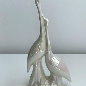 Może przedstawiać: Perłowa ceramiczna rzeźba dw&oacute;ch stylizowanych ptak&oacute;w, być może żurawi lub czapli, z długimi szyjami i eleganckimi pozami. Ptaki są splecione, co sugeruje czuły uścisk. Rzeźba ma błyszczące wykończenie i jest zamontowana na ozdobnej podstawie.