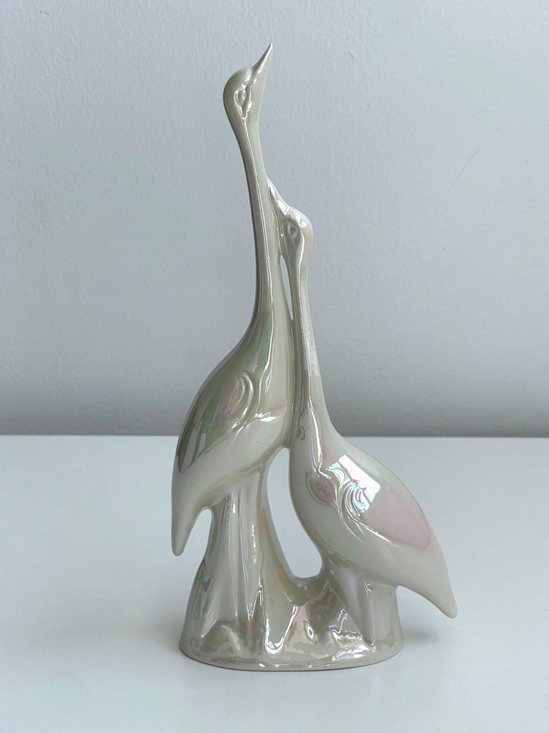 Może przedstawiać: Perłowa ceramiczna rzeźba dw&oacute;ch stylizowanych ptak&oacute;w, być może żurawi lub czapli, z długimi szyjami i eleganckimi pozami. Ptaki są splecione, co sugeruje czuły uścisk. Rzeźba ma błyszczące wykończenie i jest zamontowana na ozdobnej podstawie.