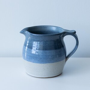 May include: A handmade ceramic jug with a blue and white glaze. The jug has a curved handle and a spout for easy pouring. The blue glaze is layered, creating a gradient effect. This decorative jug is perfect for serving beverages.