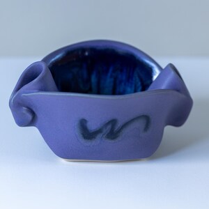 May include: A small, hand-formed ceramic bowl with a matte purple exterior and a glossy, deep blue interior. The rim features two distinct, folded-over decorative flaps on the sides, and a dark, abstract brushstroke design is centred on the front.