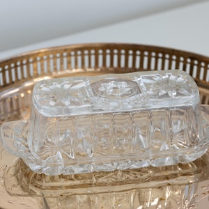 May include: A clear, rectangular glass butter dish with a starburst design on the lid and base, resting on a gold-toned metal serving tray. The tray has a decorative floral pattern and a pierced rim.