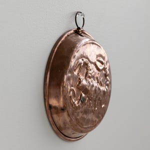 May include: A circular copper mould with an embossed design, suspended by a small metal loop. The mould is mounted on a light-coloured wall, highlighting its detailed pattern and vintage appearance.