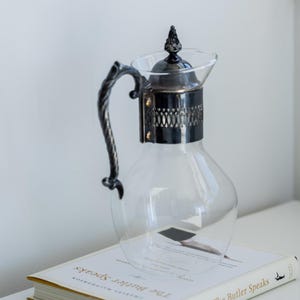 May include: A clear glass jug with a decorative metal handle and lid, sitting on a book titled "The Butler Speaks." The jug has a rounded body and a silver-coloured metal band around the neck.