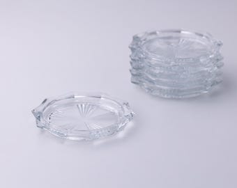 May include: A stack of six clear glass coasters with a starburst design in the center and a scalloped edge. One coaster is separate from the stack. The coasters are made of clear glass.