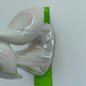 Może przedstawiać: Biała, iryzująca rzeźba ceramiczna o zakrzywionym, abstrakcyjnym wzorze. Rzeźba ma około 13 cm wysokości, mierząc zieloną linijką. Powierzchnia odbija subtelny tęczowy połysk.