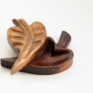 May include: A wooden leaf-shaped sculpture with a natural finish. The sculpture features a large leaf and a base with a heart-shaped bowl. The wood grain is visible, and the piece has a warm, inviting appearance.