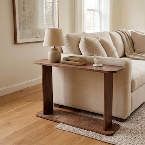 May include: A narrow, rectangular dark wood console table with rounded edges, featuring a flat top surface supported by two vertical side panels and a matching base plate. The table is positioned horizontally, providing a stable surface for items.