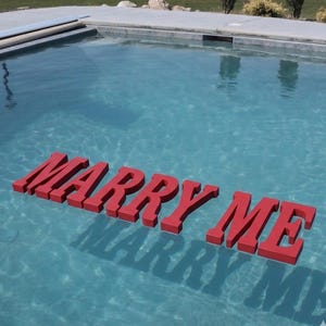 May include: Red floating letters spelling "MARRY ME" in a swimming pool. The letters cast a shadow on the water. The pool has a light blue color and is surrounded by a concrete deck.