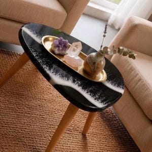 May include: A teardrop-shaped side table with a black and white resin top and three light wood legs. On the table sits an oval gold tray containing a purple amethyst cluster, a clear quartz point, a rose quartz stone, and a small ceramic vase.